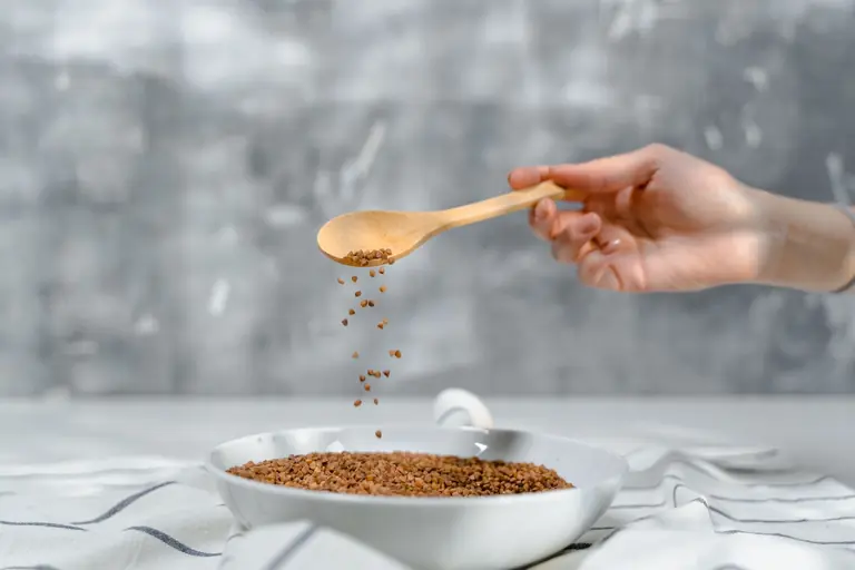 A close-up of a hand pouring flaxseeds into a bowl using a wooden spoon.