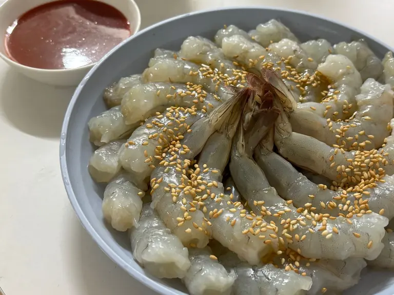 Plate of raw shrimp garnished with sesame seeds and a spicy dipping sauce.