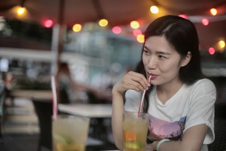 Asian woman enjoying a cocktail in a cozy outdoor setting with ambient lights.