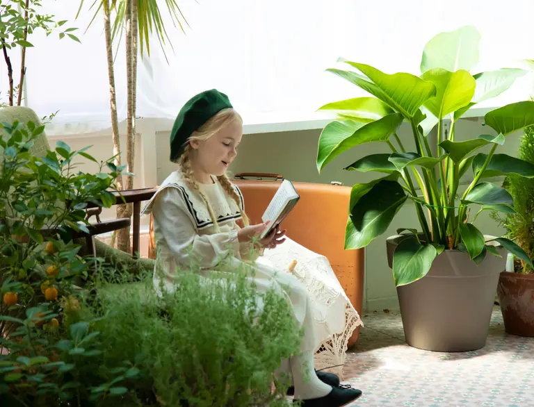 A young girl with braided hair reads a book surrounded by lush houseplants indoors.
