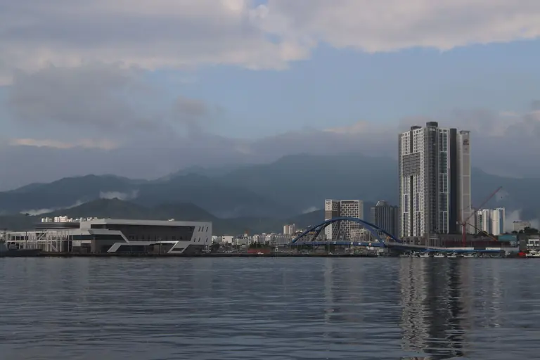 Cityscape featuring modern architecture, a blue bridge, and mountain range under cloudy sky.
